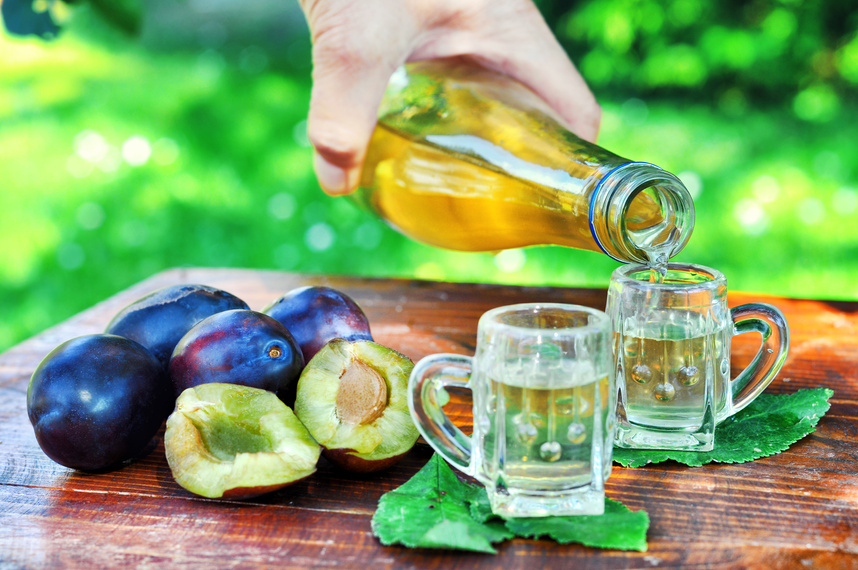 A well-known brandy rakia slivovitz in shot glasses with plums on wooden table in orchard. Traditional Serbian brand alcoholic drink of organic production.Human hand pouring a well-known brandy rakia